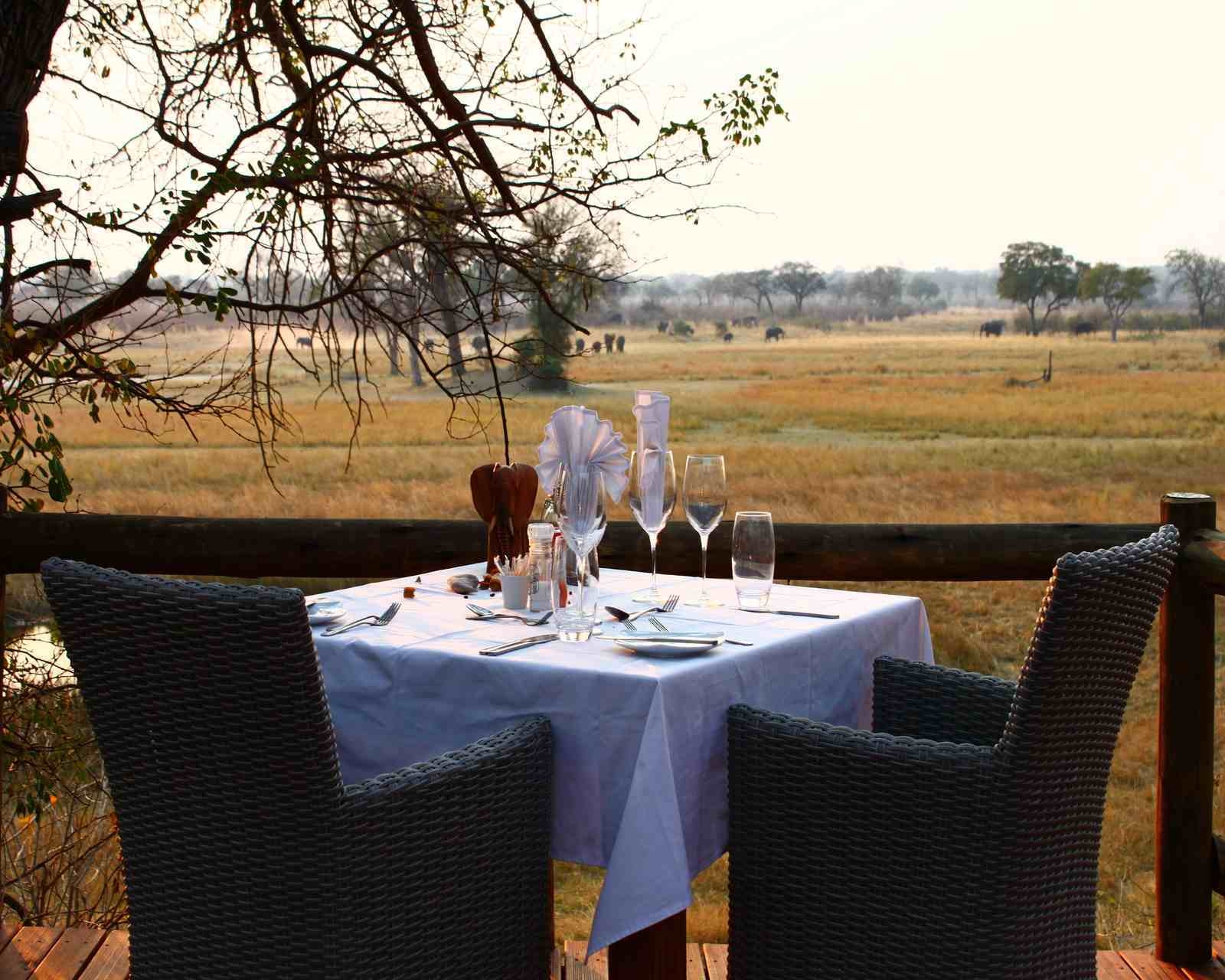 A table is laid with a white tablecloth, cutlery and glasses, on a deck overlooking the plains where many elephants can be seen in the distance.