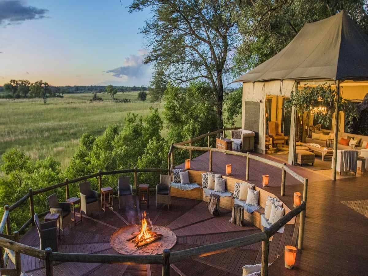 A large wooden deck with an open-sided tent with sofas and low lighting, and a large, circular fire pit area with seating, overlooking the floodplains