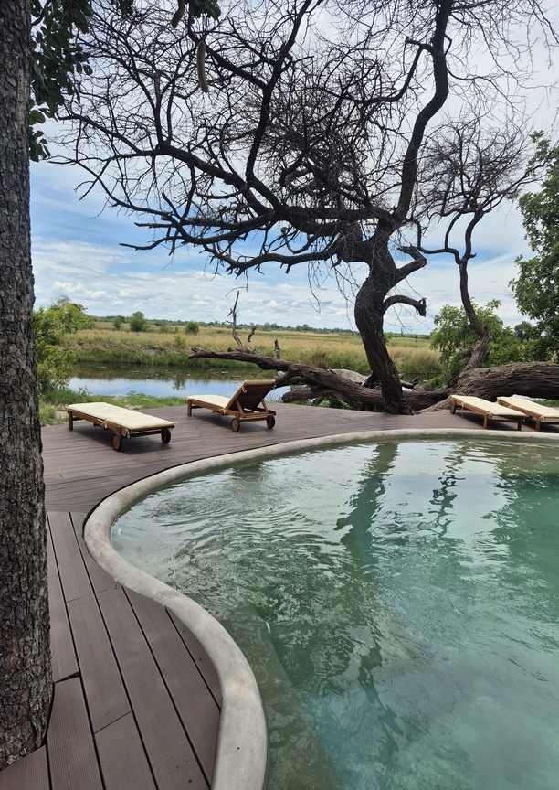 The curved edge of a swimming pool is in the foreground, with four wooden sun loungers on a wooden deck that overlooks the river.