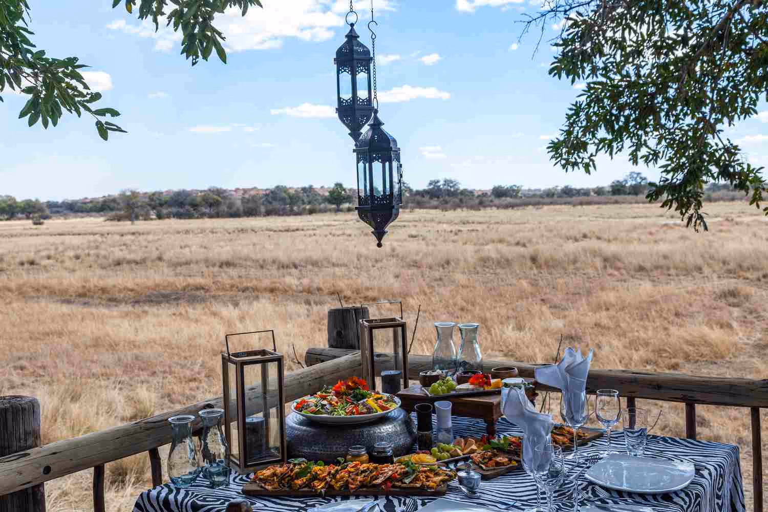 A table set with a black and white tablecloth, food and wine glasses looks over the plains, framed by trees and with two lanterns above.