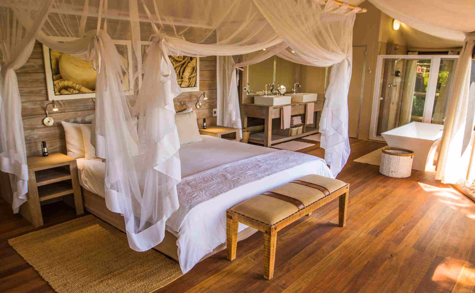 A double bed is surrounded by white drapes in a suite with wooden floors, wood-panelled walls, art on the walls and a bathroom area with two sinks and a large bathtub in the background.