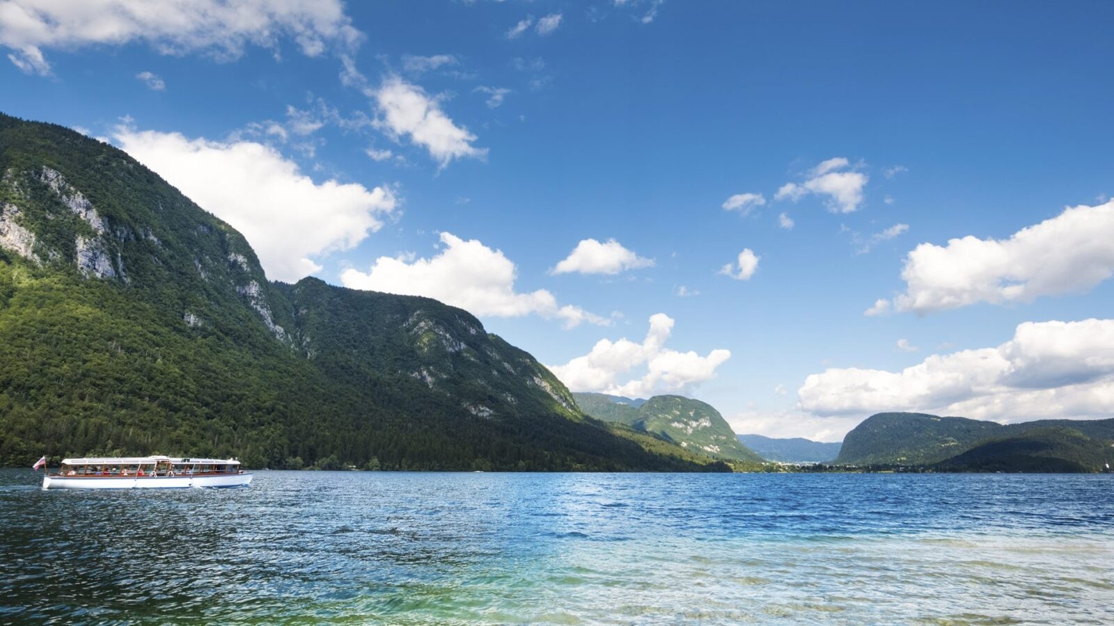 View over large clear Lake Bohinj with small white sightseeing boat motoring across the water. The lake is surrounded by forested mountains.