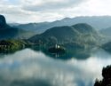 Aerial view of Lake Bled in the Alps, with a small church on an island in the middle of clear still lake surrounded by mountains and thick forests