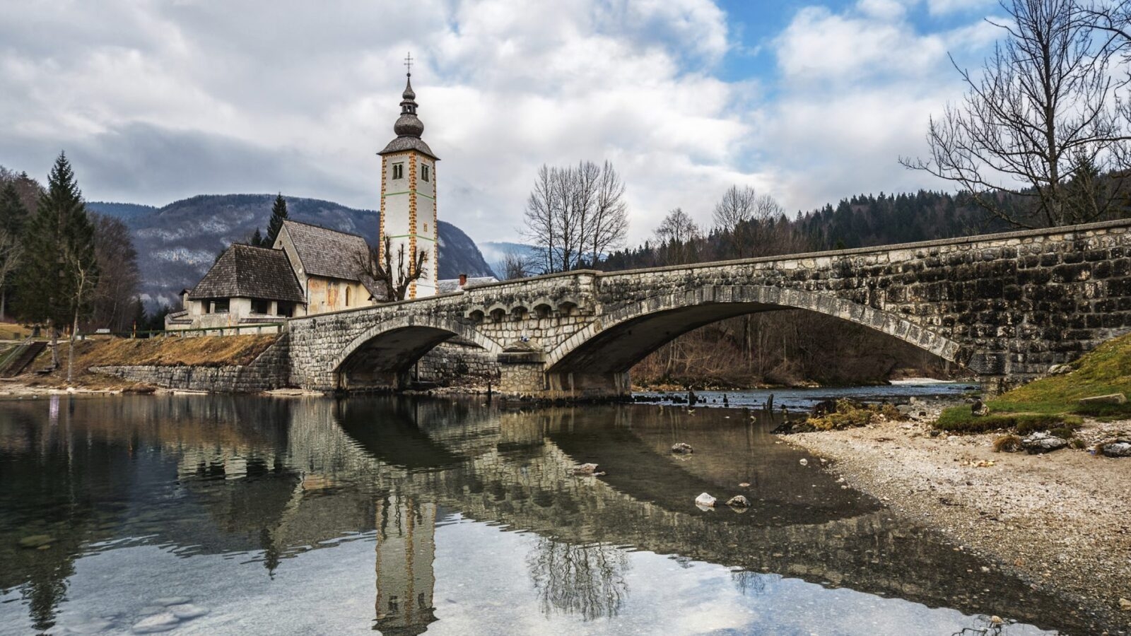 A small church by an arched stone bridge over a shallow river in Slovenia