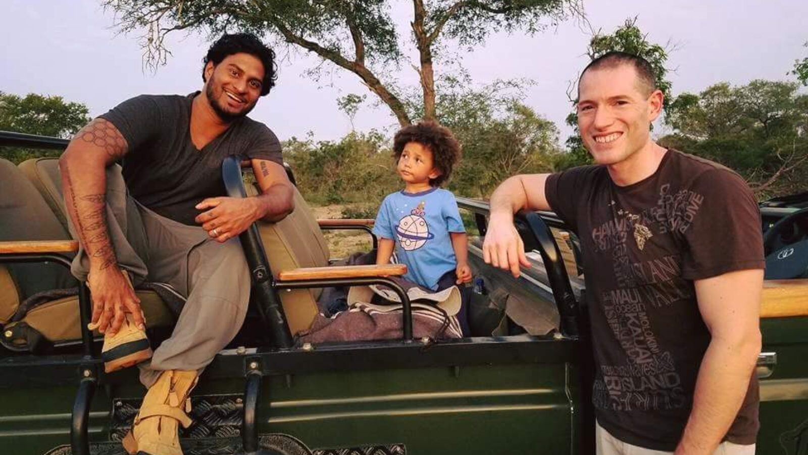 Two men and a child sitting in an open safari vehicle with trees in the background.
