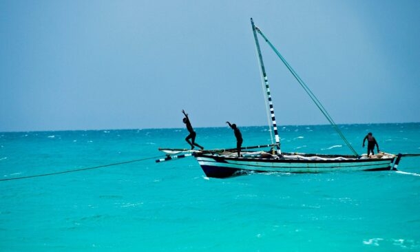 People jumping into the ocean from a traditional wooden boat with a single sail on a clear day.
