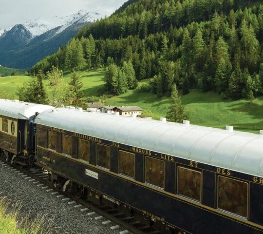 The Venice Simplon Orient Express travelling through the lush green countryside bordered by snowcapped mountains