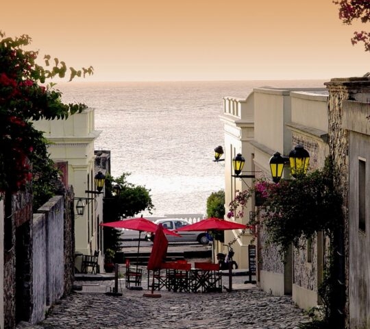 Cobblestone street with red umbrellas and vintage buildings leading toward the sea under a warm sunset sky.