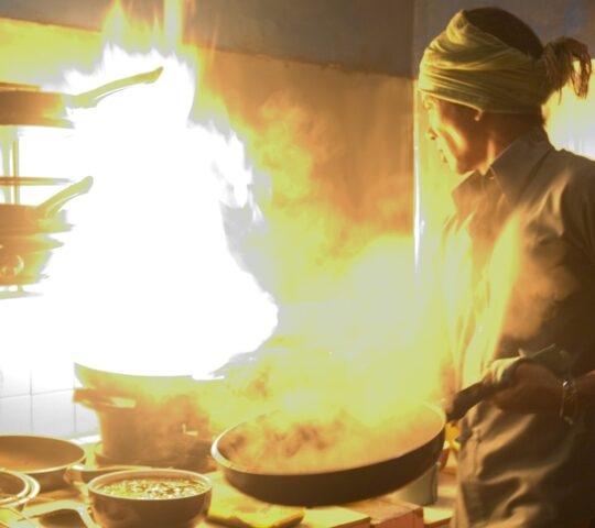 A person cooking with a wok as bright yellow flames rise high into the air in a kitchen setting.