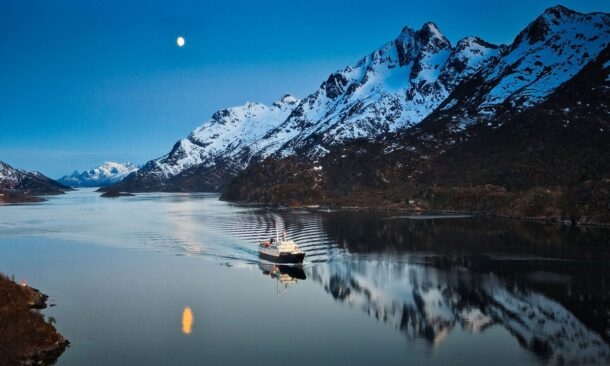 A ship sailing through a calm fjord at night with snowy mountains and a full moon reflected in the water.