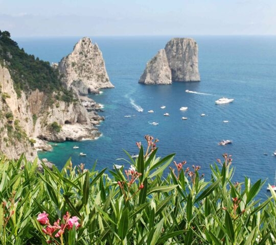 A high-angle coastal view showing two large rock stacks in the ocean with boats scattered nearby.