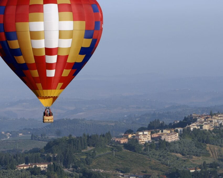 A patterned hot air balloon flies above a hazy green valley featuring traditional Italian stone buildings and trees.