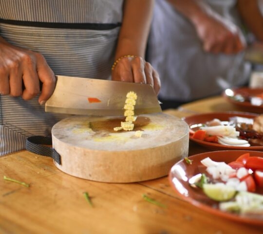 Close-up of hands using a metal cleaver to slice cloves of garlic on a round wooden block.