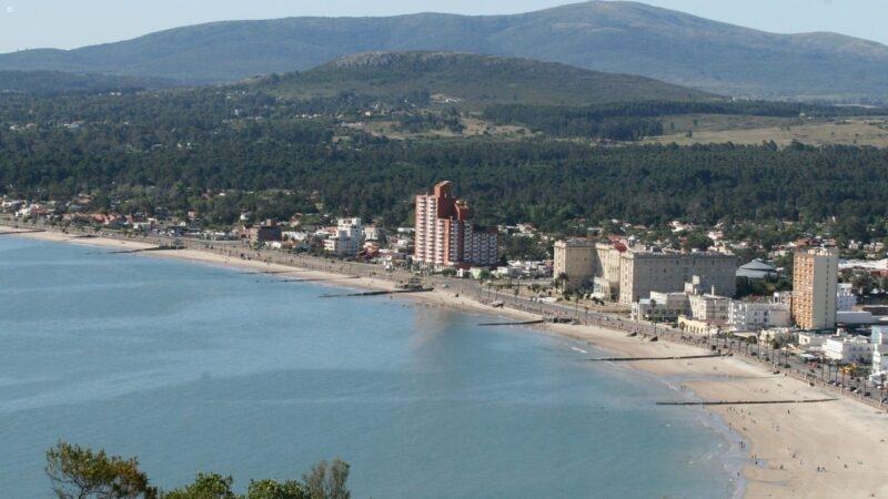 High-angle panorama of a city coastline and sandy beach with a large mountain under a clear blue sky.
