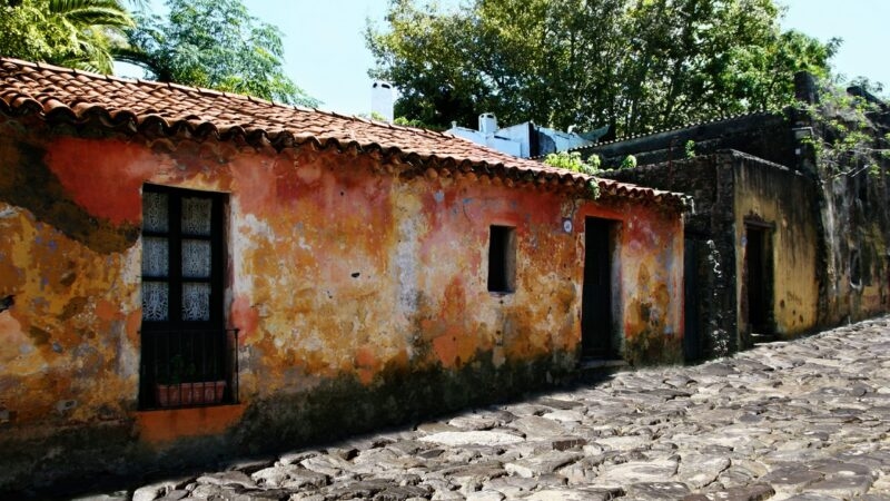 Rustic house with colorful weathered walls and a brown tiled roof on a historic cobblestone street.