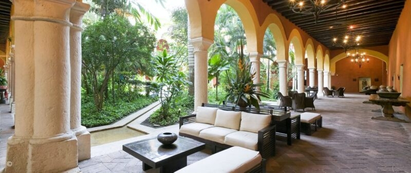 An elegant outdoor courtyard features stone arches, white sofas, and lush tropical greenery in a central garden.