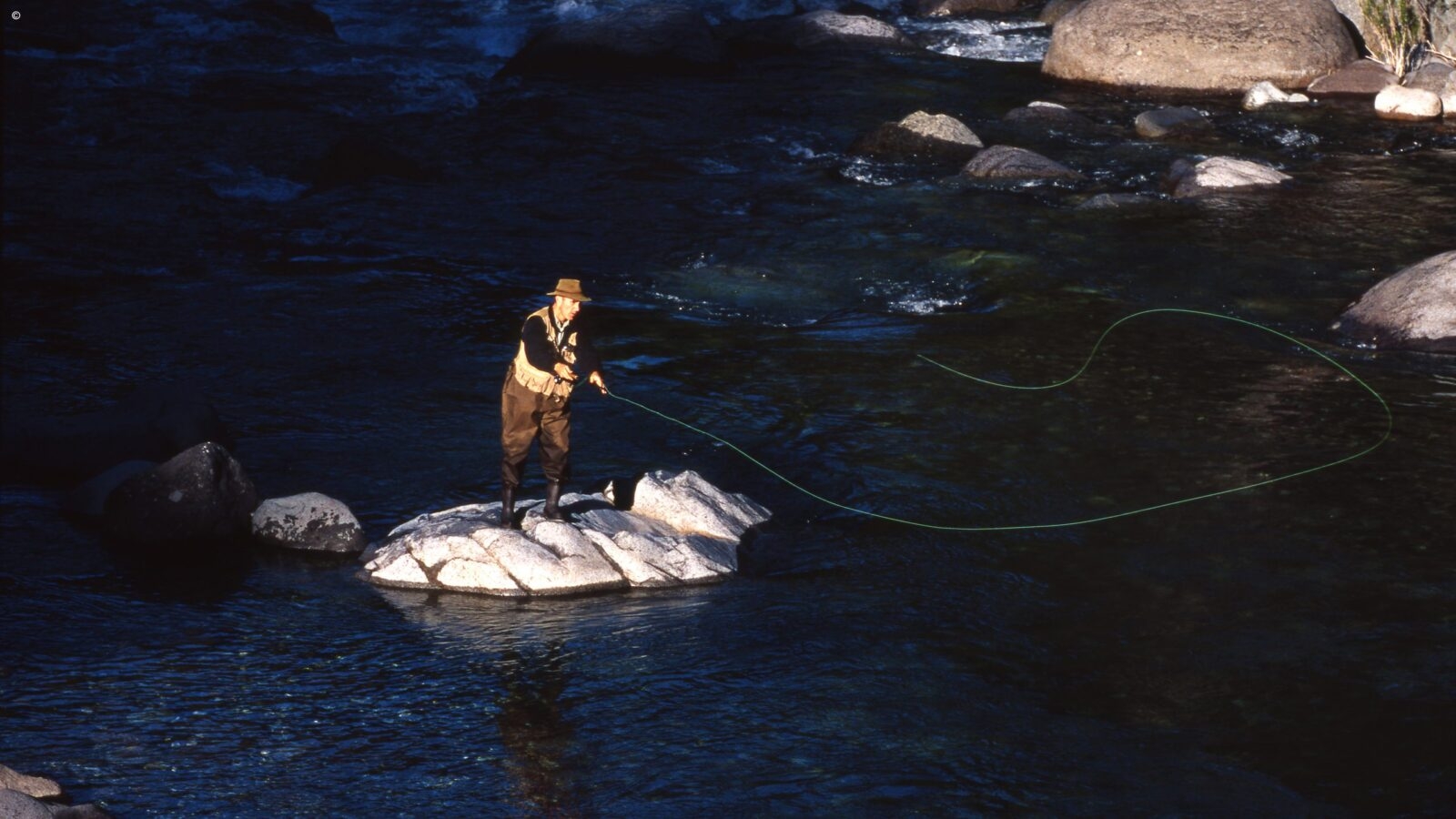 A man in brown trousers and vest and a brown brimmed hat standing on a rock in a river casting a long fishing line into the water