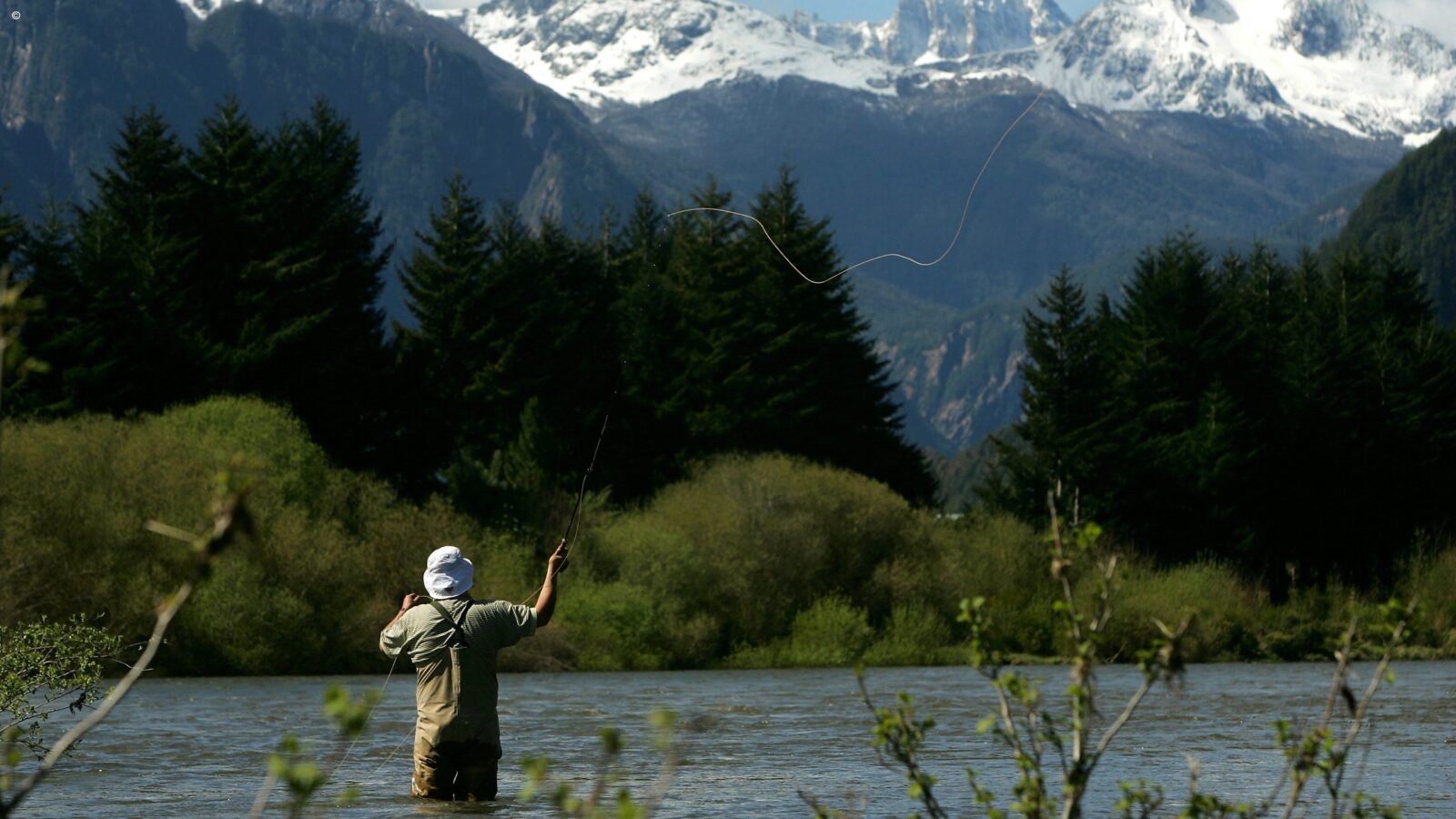 A man in green overalls and a white bucket hat standing in a lake casting a fishing line into the water. There are snow capped mountains and foliage in the background