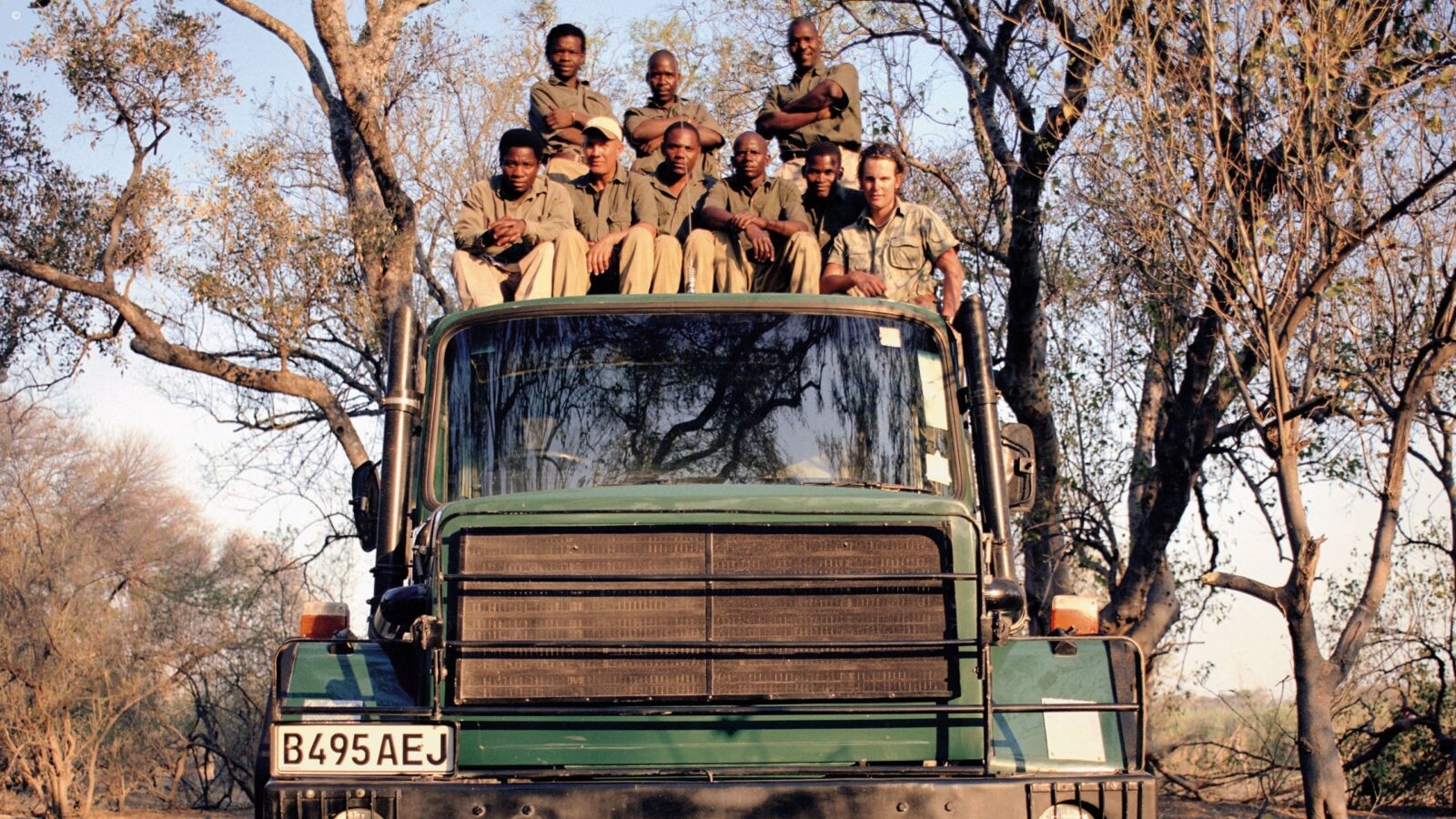 Guides in green shorts and sand-coloured trousers sitting on top of a large green safari truck beneath a tree