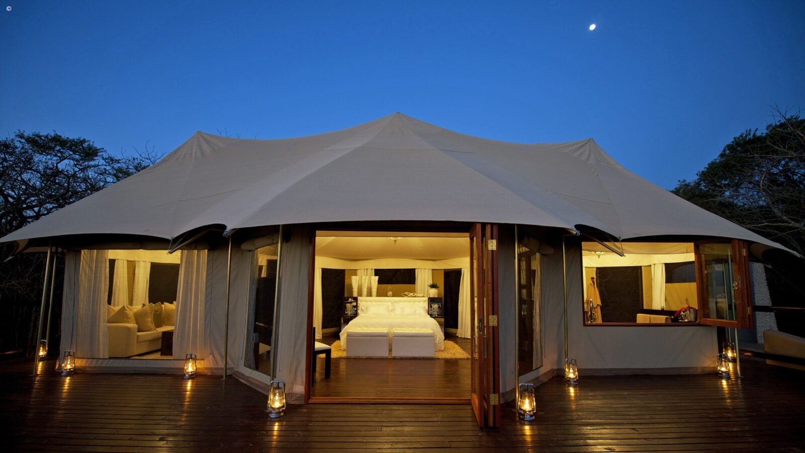 Large luxury safari tent illuminated at night under a crescent moon for honeymoon grand tours to Africa.