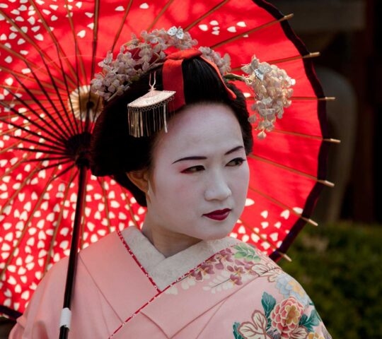 Side view of a woman in a traditional kimono and geisha makeup walking past historic wooden buildings.