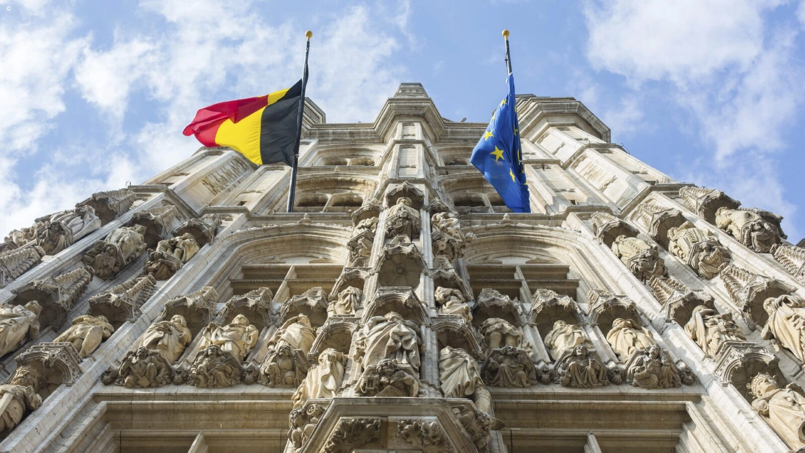 Shot looking up the front of a tall historic building with plae stone walls, detailed stone carvings of people, and the Belgian and European flags flying from two windows