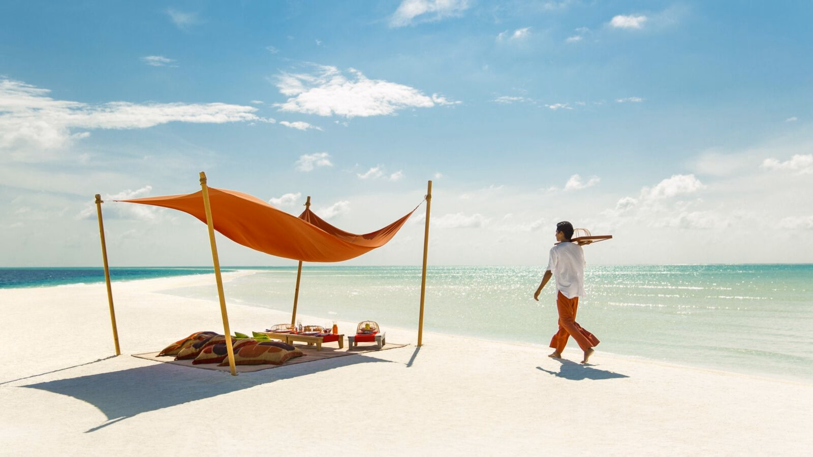 A mean carrying a tray towards a picnic setup under a shelter on a white sandy beach by the sea