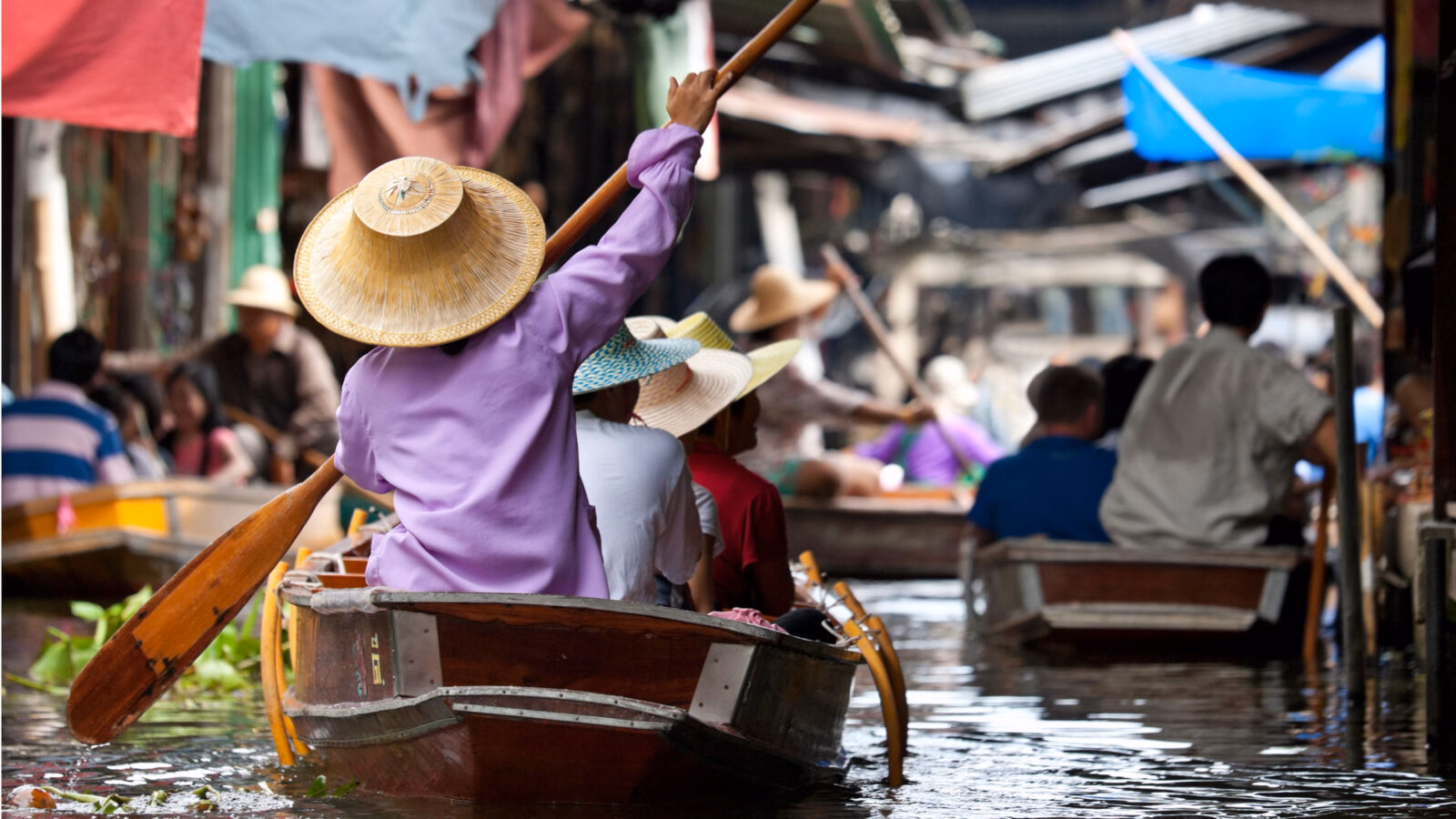 People wearing traditional hats row wooden boats through a busy floating market canal.