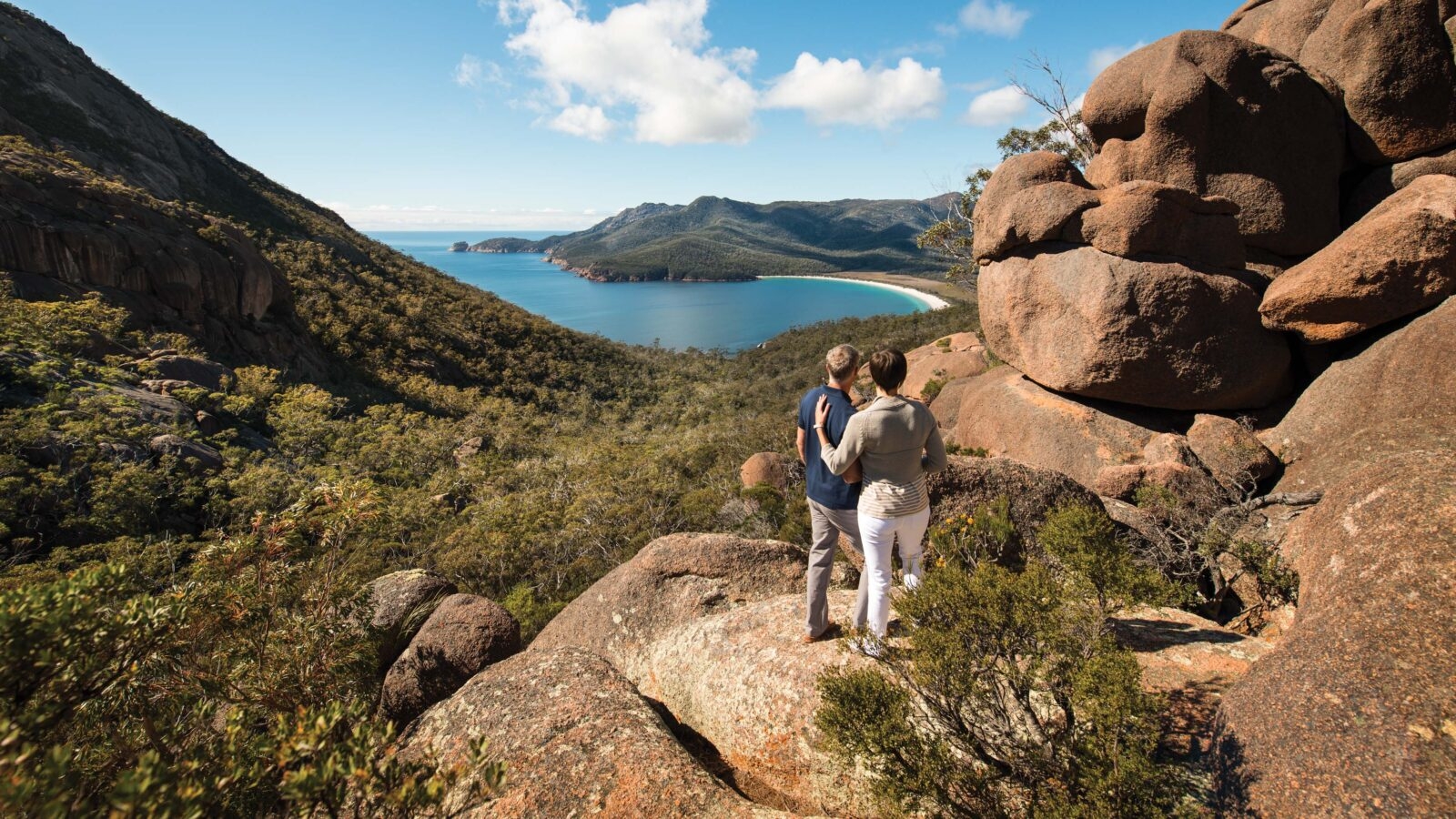 Two people standing at a scenic lookout point on a rocky hill overlooking a beautiful ccove with a white sandy beach and blue sea
