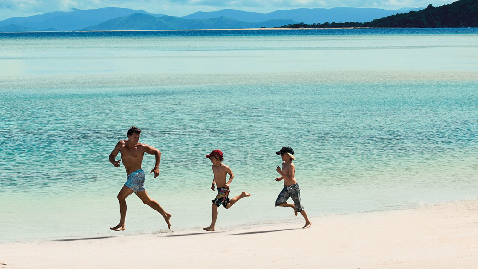 A man and two children running on a white sand beach next to clear turquoise water.