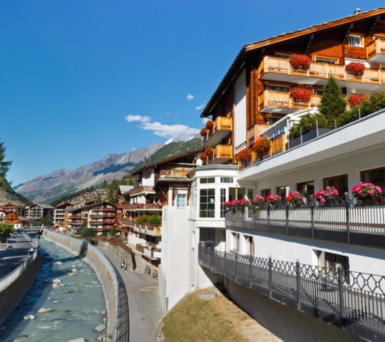Large wooden buildings with red flowers on balconies situated next to a canal and mountains.