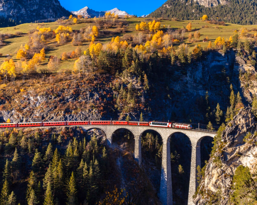 Panoramic view of a red train on a stone viaduct bridge surrounded by autumn foliage and mountains.