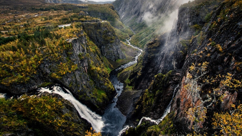 A dramatic deep canyon with a winding river and waterfall surrounded by steep, mossy rock walls and mist.