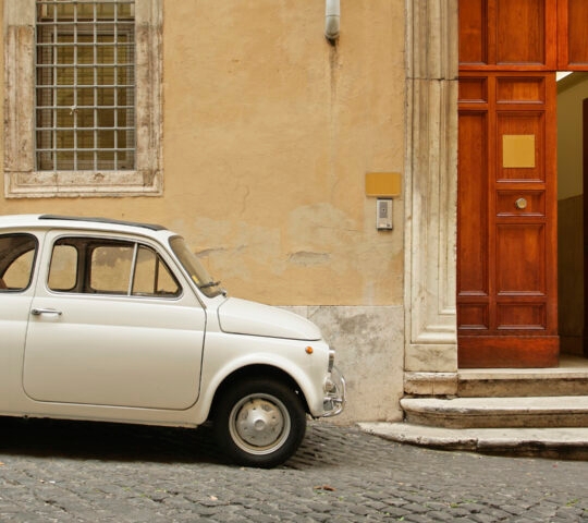 A small white vintage car parked on a sloping cobblestone street next to a building with a tall wooden doorway.