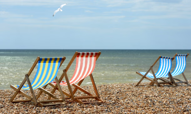 Yellow, red, and blue striped beach chairs on a shingle shore overlooking the ocean with a bird in flight.