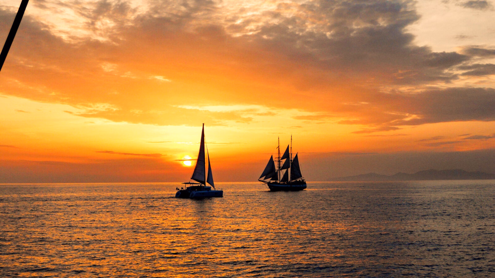 Silhouette of two sailboats on the sea during a vibrant orange and yellow sunset with scattered clouds.