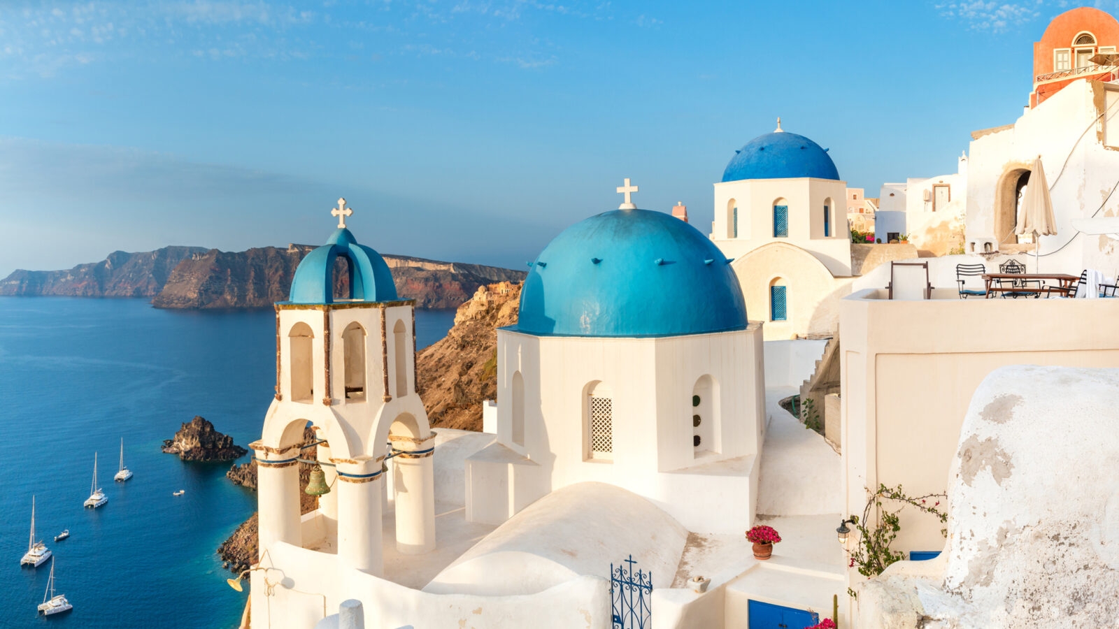 White churches with bright blue domes on a cliffside overlooking a deep blue sea under a clear sky.
