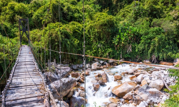 Narrow wooden suspension bridge crossing over a white-water rocky river into a lush green forest.