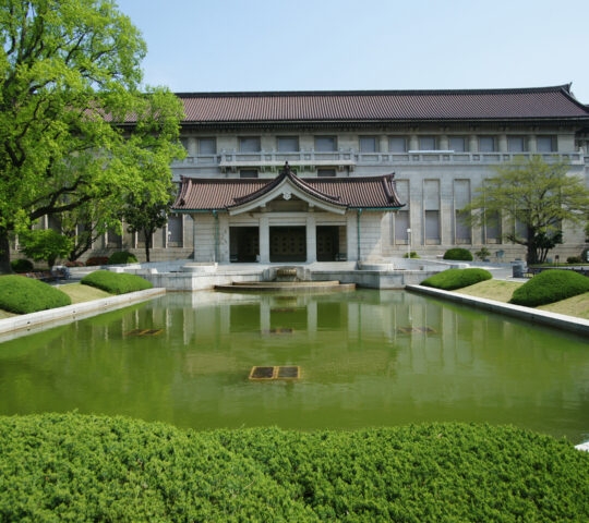 Large traditional museum building with a rectangular reflecting pond and shaped green bushes in front.