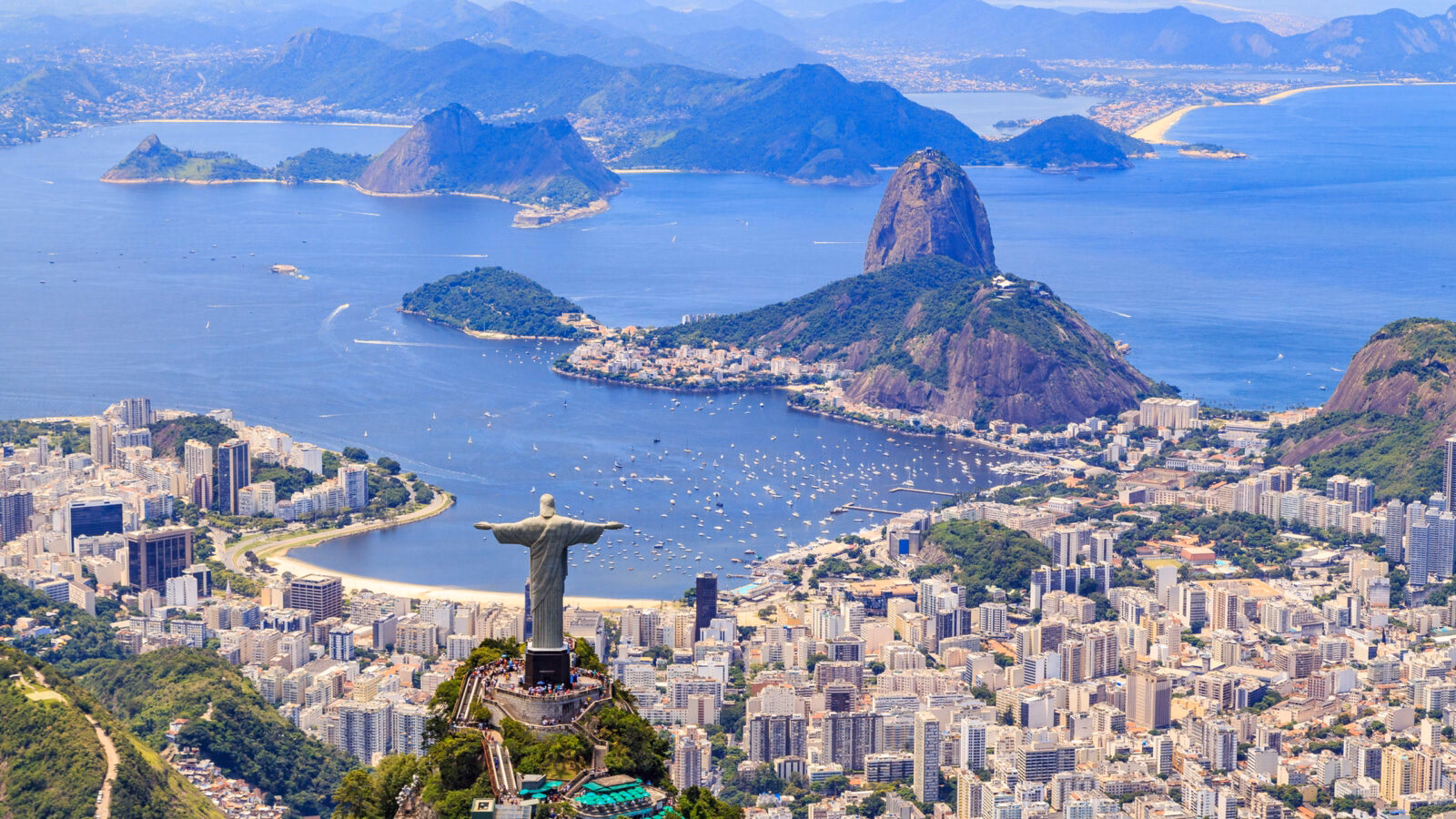 The Christ the Redeemer statue stands on a mountain peak overlooking a coastal city and bay.