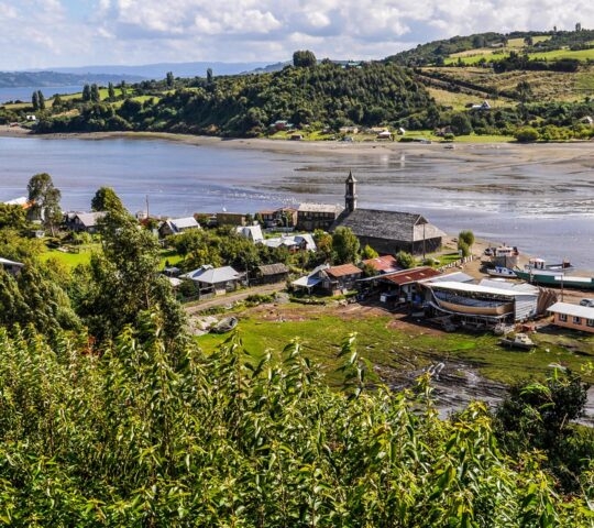 A small coastal village with a dark church is nestled in a green valley by the water, surrounded by hills.
