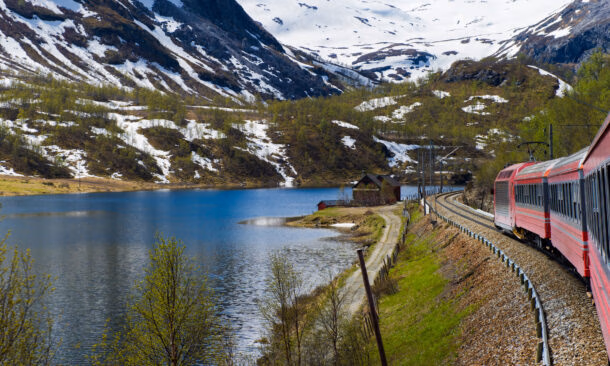 A red train traveling on tracks alongside a blue lake with snow-capped mountains in the background.