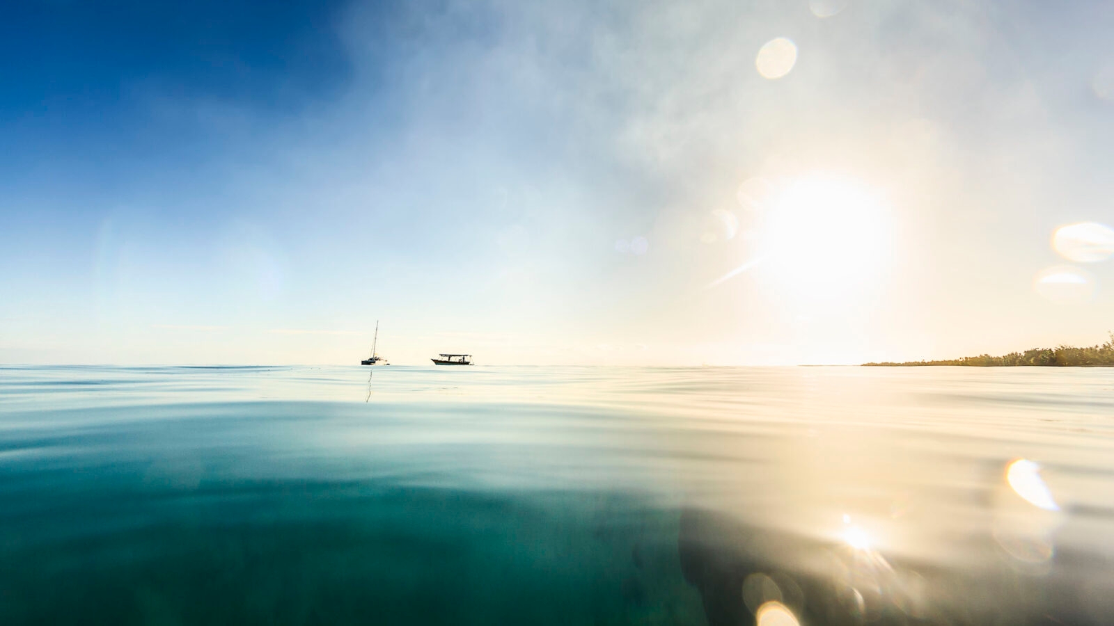 A DSLR photo taken from inside the sea, just above surface level, in tranquil water in Rangiroa, French Polynesia. The sun is low on the horizon and will bet set in a few moments. It is produncing some flare in the water drops on the lens. Two boats are anchored to the left of the frame.
