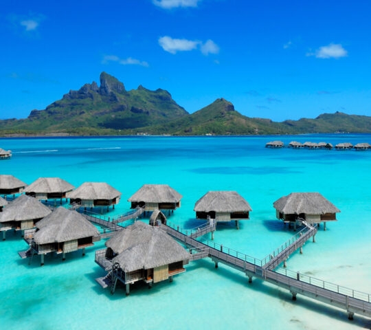 Thatched roof overwater bungalows over clear turquoise water on luxury French Polynesia trips.