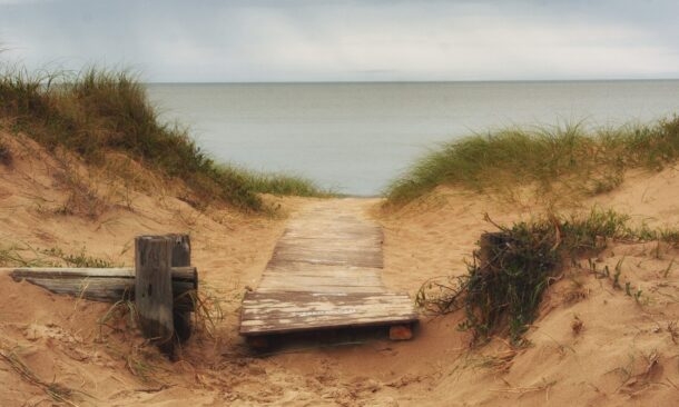 Rustic wooden path through sand dunes leading to the ocean under a cloudy sky.