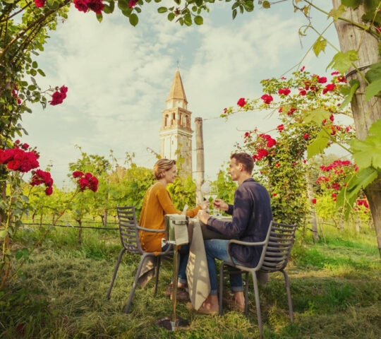 A man and woman dining outdoors in a garden surrounded by red rose bushes and a tall historic tower.