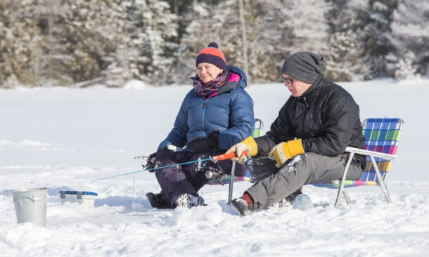 A man and woman in winter coats and hats sit on folding chairs ice fishing on a wide snowy plain.