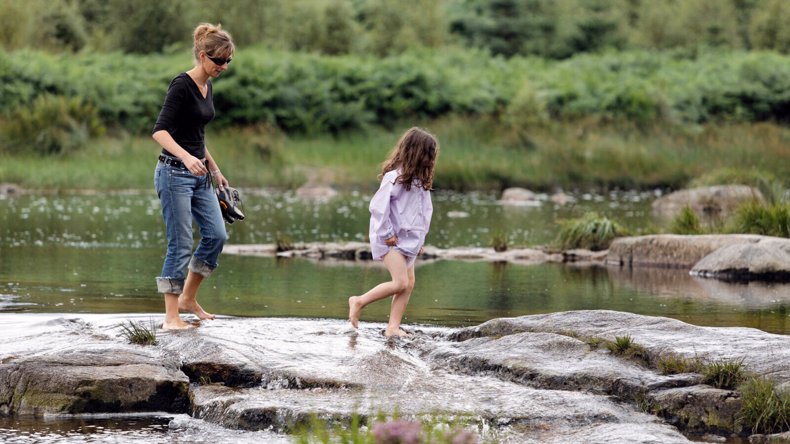 A woman and a young girl wade across a rocky stream in a green woodland setting.