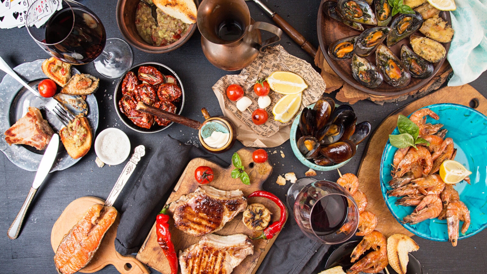 Large table spread with grilled meat, mussels in shells, cooked shrimp, salmon, and glasses of red wine.