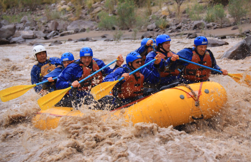 A group of eight people white-water rafting on a turbulent brown river, paddling a yellow inflatable boat. Luxury Argentina holidays.